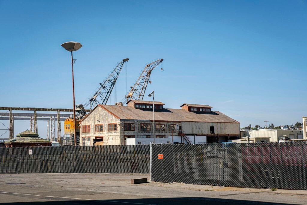 Rustic industrial building with cranes against a clear blue sky, fenced area, daytime.